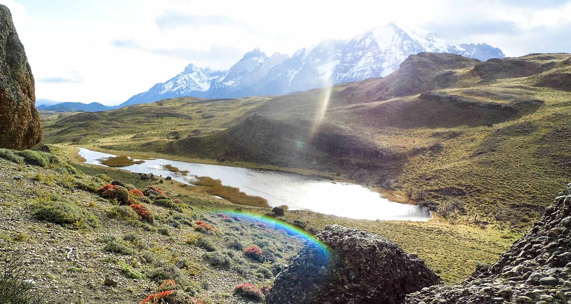 Patagonian steppe and a lake below snowy Andes peaks, with a rainbow over the grassland